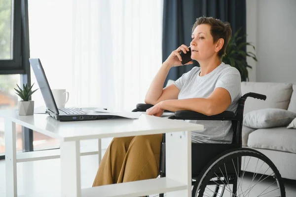 Freelancer in wheelchair using laptop near notebook and papers on table.