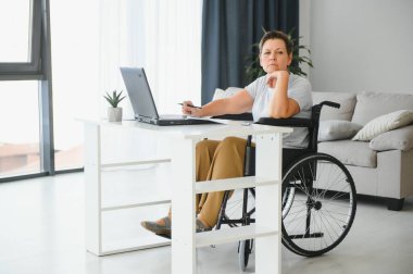 Tekerlekli sandalyedeki serbest yazar dizüstü bilgisayarını ve masadaki kağıtları kullanıyor..