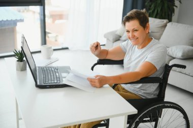 Tekerlekli sandalyedeki serbest yazar dizüstü bilgisayarını ve masadaki kağıtları kullanıyor..