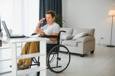Tekerlekli sandalyedeki serbest yazar dizüstü bilgisayarını ve masadaki kağıtları kullanıyor..