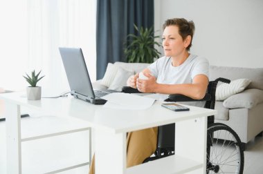 Tekerlekli sandalyedeki serbest yazar dizüstü bilgisayarını ve masadaki kağıtları kullanıyor..