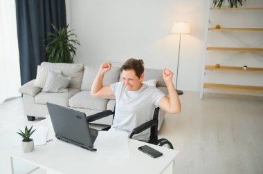 Evde tekerlekli sandalyede oturan orta yaşlı kadın dizüstü bilgisayar kullanıyor..