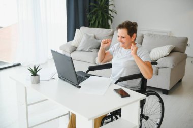 Evde tekerlekli sandalyede oturan orta yaşlı kadın dizüstü bilgisayar kullanıyor..