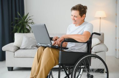 Evde tekerlekli sandalyede oturan orta yaşlı kadın dizüstü bilgisayar kullanıyor..