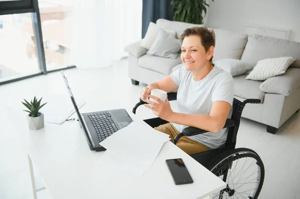 Freelancer in wheelchair using laptop near notebook and papers on table.