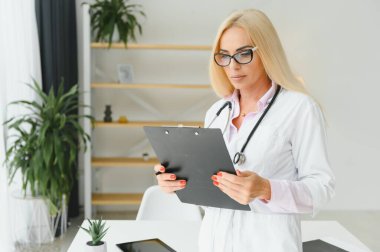 Laboratuvar önlüğü ve steteskop giyen kadın doktor hastanede dikilirken elinde pano tutuyormuş.