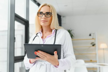 Elinde dijital tablet tutan güzel olgun kadın doktor portresi. Dijital tablet kullanan kendine güvenen kadın doktor ve hastanede arka planda konuşan meslektaşı.