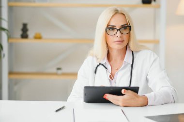 Orta yaşlı kadın doktorun portre fotoğrafı. Masada oturuyor ve doktor ofisinde çalışıyor..