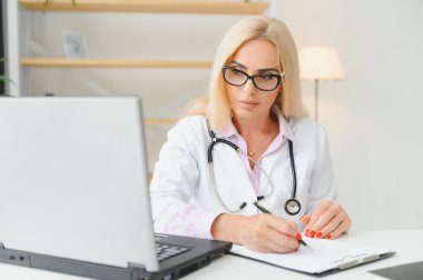 Orta yaşlı kadın doktorun portre fotoğrafı. Masada oturuyor ve doktor ofisinde çalışıyor..