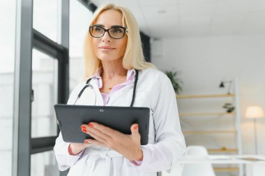 Elinde dijital tablet tutan güzel olgun kadın doktor portresi. Dijital tablet kullanan kendine güvenen kadın doktor ve hastanede arka planda konuşan meslektaşı.