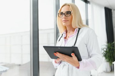 Elinde dijital tablet tutan güzel olgun kadın doktor portresi. Dijital tablet kullanan kendine güvenen kadın doktor ve hastanede arka planda konuşan meslektaşı.