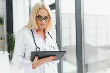 Elinde dijital tablet tutan güzel olgun kadın doktor portresi. Dijital tablet kullanan kendine güvenen kadın doktor ve hastanede arka planda konuşan meslektaşı.
