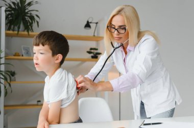 Pediyatrist nefes alışını ve kalp atışlarını steteskopla dinliyor. Doktoru ziyaret eden sevimli küçük çocuğun portresi.