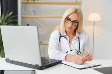 Orta yaşlı kadın doktorun portre fotoğrafı. Masada oturuyor ve doktor ofisinde çalışıyor..