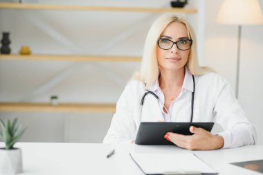 Orta yaşlı kadın doktorun portre fotoğrafı. Masada oturuyor ve doktor ofisinde çalışıyor..