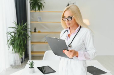 Orta yaşlı kadın doktorun portre fotoğrafı. Masada oturuyor ve doktor ofisinde çalışıyor..