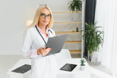 Orta yaşlı kadın doktorun portre fotoğrafı. Masada oturuyor ve doktor ofisinde çalışıyor..