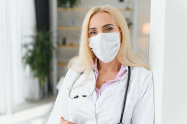 Klinikte duran, doktor üniforması ve tıbbi maske takan orta yaşlı sarışın kadın..
