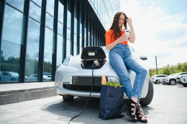 Beklerken akıllı telefon kullanmak. Elektrikli arabalardaki kadın gündüz şarj istasyonunda. Yepyeni bir araç.