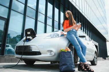 Beklerken akıllı telefon kullanmak. Elektrikli arabalardaki kadın gündüz şarj istasyonunda. Yepyeni bir araç.
