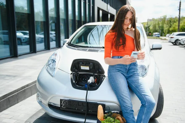 Young woman is standing near the electric car and looks at the smart phone. The rental car is charging at the charging station for electric vehicles. Car sharing