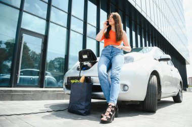 Genç kadın elektrikli arabanın yanında duruyor ve akıllı telefona bakıyor. Kiralık araba elektrikli araçlar için şarj istasyonunda şarj oluyor. Araba paylaşımı