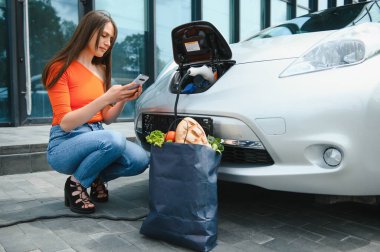Genç kadın elektrikli arabanın yanında duruyor ve akıllı telefona bakıyor. Kiralık araba elektrikli araçlar için şarj istasyonunda şarj oluyor. Araba paylaşımı
