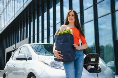 Genç kadın elektrikli arabanın yanında duruyor. Kiralık araba elektrikli araçlar için şarj istasyonunda şarj oluyor. Araba paylaşımı