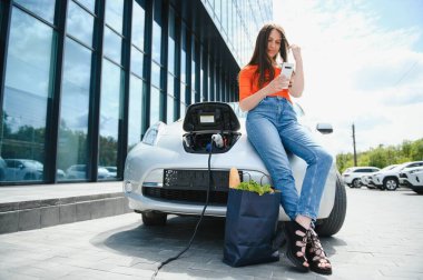 Genç kadın elektrikli arabanın yanında duruyor ve akıllı telefona bakıyor. Kiralık araba elektrikli araçlar için şarj istasyonunda şarj oluyor. Araba paylaşımı