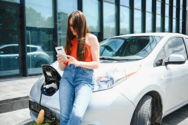 Genç kadın elektrikli arabanın yanında duruyor ve akıllı telefona bakıyor. Kiralık araba elektrikli araçlar için şarj istasyonunda şarj oluyor. Araba paylaşımı