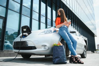 Genç kadın elektrikli arabanın yanında duruyor. Kiralık araba elektrikli araçlar için şarj istasyonunda şarj oluyor. Araba paylaşımı