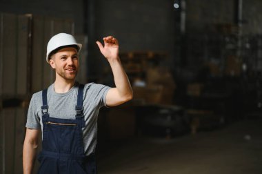 Depoda rafların arasında duran beyaz kasklı mutlu erkek işçinin portresi.