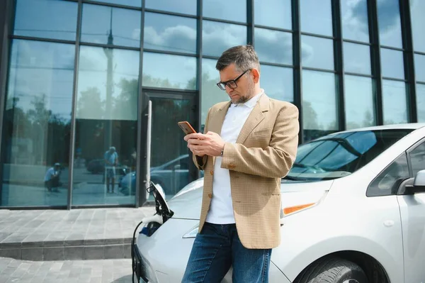 The guy sat down on the hood of the car. His car is charging at the charging station. A man looks at the smartphone screen and smiles