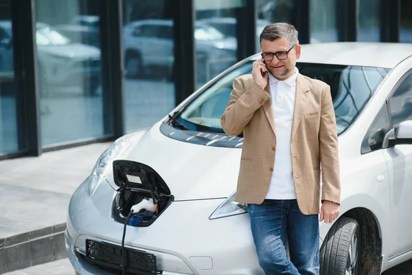 The guy sat down on the hood of the car. His car is charging at the charging station. A man looks at the smartphone screen and smiles