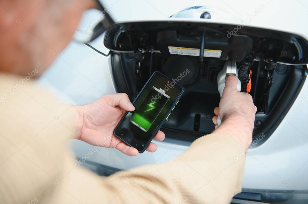 Man Charging Electric Vehicle With Cable Looking At App On Mobile Phone.