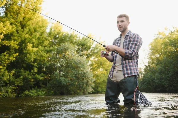 Man with fishing rod, fisherman men in river water outdoor. Catching trout fish in net. Summer fishing hobby.