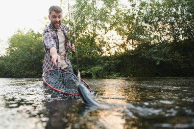 Man with fishing rod, fisherman men in river water outdoor. Catching trout fish in net. Summer fishing hobby.
