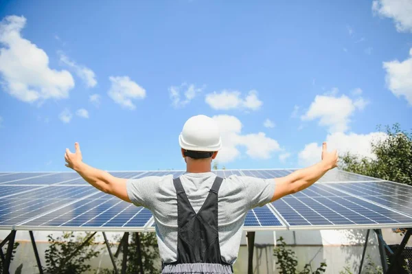 Smiling worker with solar station, raising his hands, showing thumbs up on a background of photovoltaic panels near the house. Man in orange uniform. Science solar energy. Renewable energy.