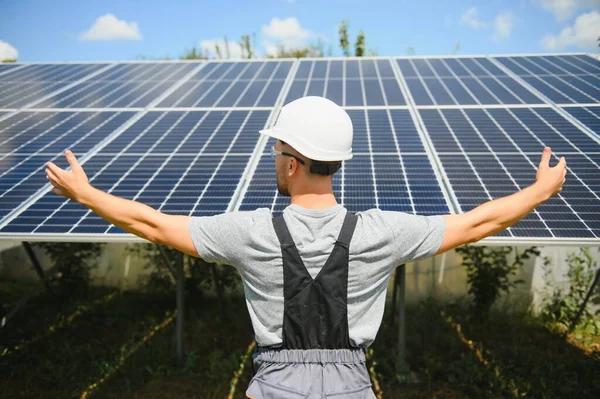 Smiling worker with solar station, raising his hands, showing thumbs up on a background of photovoltaic panels near the house. Man in orange uniform. Science solar energy. Renewable energy.