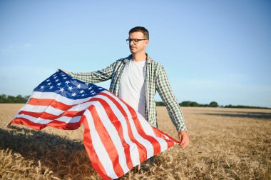 Young patriotic farmer stands among new harvest. Boy walking with the american flag on the wheat field celebrating national independence day. 4th of July concept