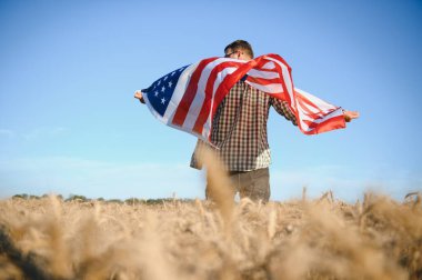 Young patriotic farmer stands among new harvest. Boy walking with the american flag on the wheat field celebrating national independence day. 4th of July concept