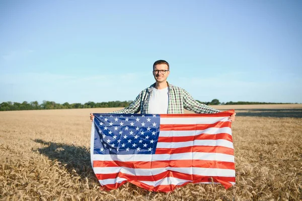 Young patriotic farmer stands among new harvest. Boy walking with the american flag on the wheat field celebrating national independence day. 4th of July concept