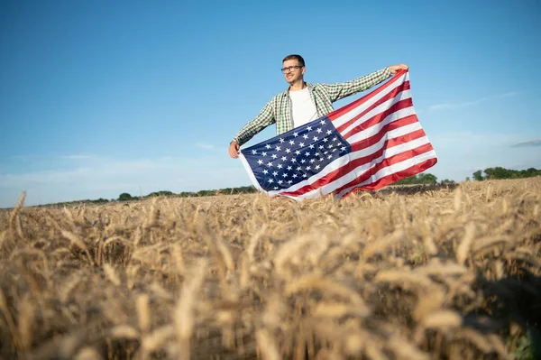 Young patriotic farmer stands among new harvest. Boy walking with the american flag on the wheat field celebrating national independence day. 4th of July concept