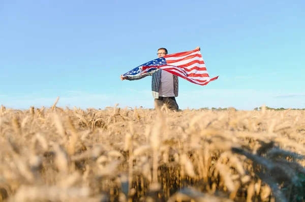 Young patriotic farmer stands among new harvest. Boy walking with the american flag on the wheat field celebrating national independence day. 4th of July concept