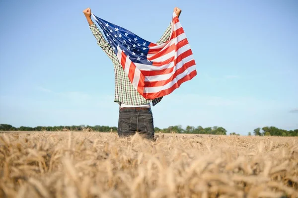 Young patriotic farmer stands among new harvest. Boy walking with the american flag on the wheat field celebrating national independence day. 4th of July concept