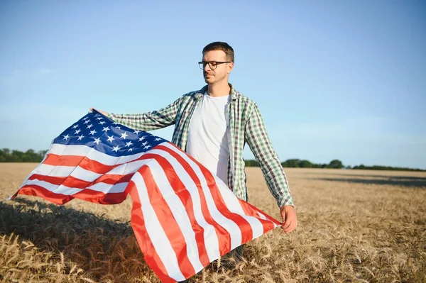 Young patriotic farmer stands among new harvest. Boy walking with the american flag on the wheat field celebrating national independence day. 4th of July concept