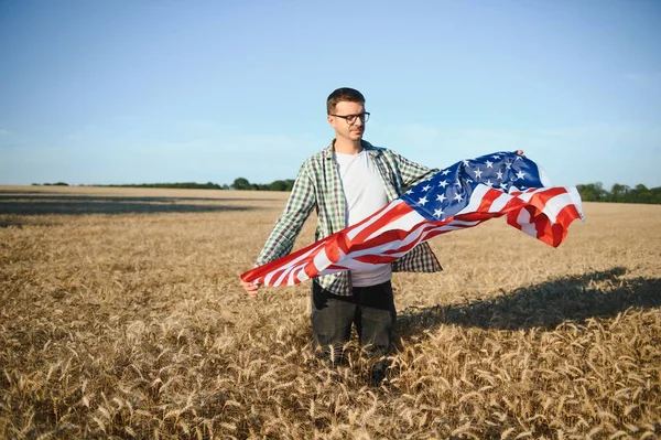 Young patriotic farmer stands among new harvest. Boy walking with the american flag on the wheat field celebrating national independence day. 4th of July concept