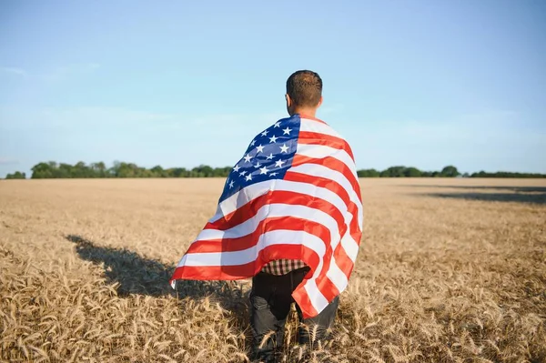 Young patriotic farmer stands among new harvest. Boy walking with the american flag on the wheat field celebrating national independence day. 4th of July concept