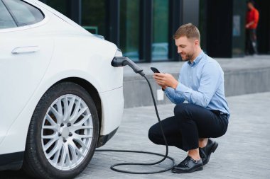 Man charging his electric car at charge station and using smartphone.