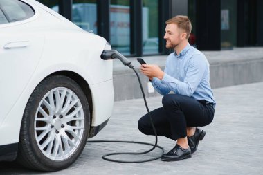 Yakışıklı sakallı adam yeni modern elektrikli arabasının yanında oturuyor ve şarj aletinin fişini çekiyor, araba şarj istasyonunda şarj olurken